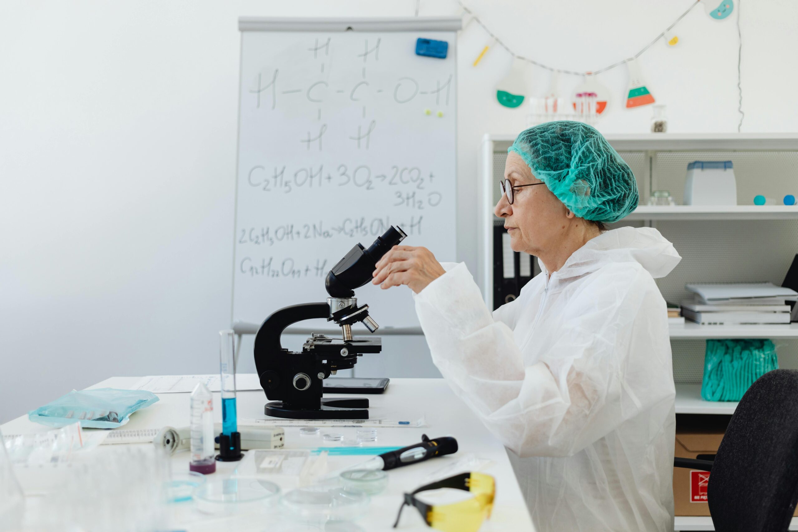 Senior scientist using a microscope in a laboratory setting wearing protective gear.